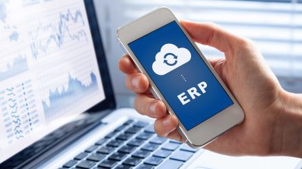 Eine Person hält ein Mobiltelefon mit einem Cloud- und ERP-Symbol in der Hand. Im Hintergrund ist ein PC-Bildschirm mit Dashboards zu sehen.