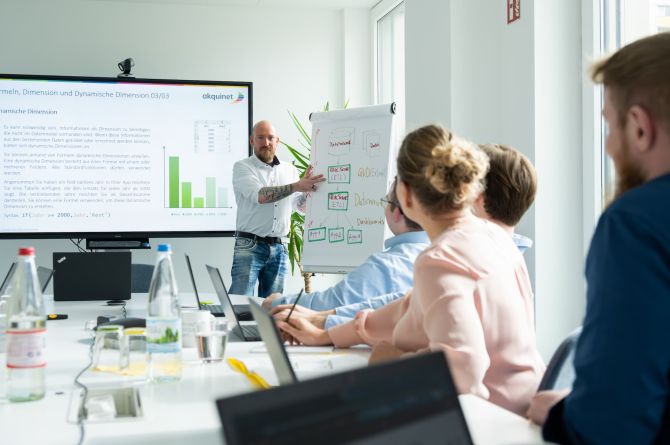 Schulungsteilnehmer mit Laptops am Tisch, der Trainer zeigt was am Flipchart