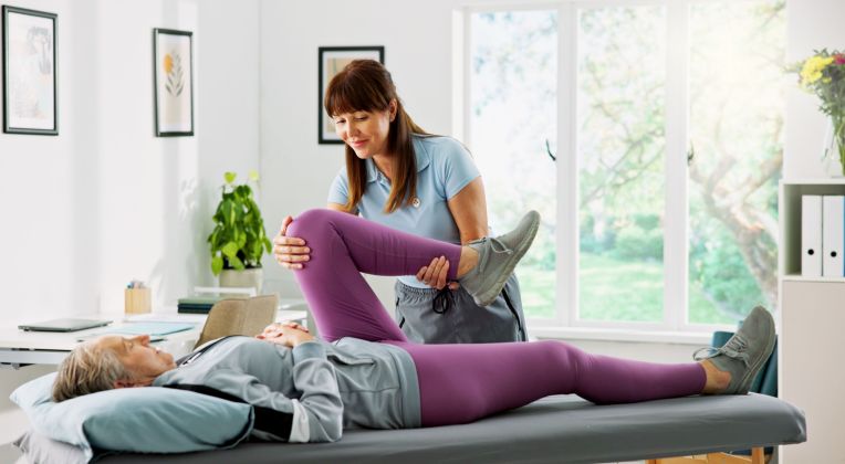 Eine Physiotherapeutin hilft einer älteren Frau auf einer Liege Übungen zu machen