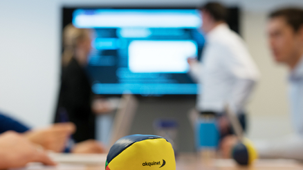 Im Hintergrund diskutieren zwei Microsoft Kollegen vor einem Fernseher. Vor der Kameralinse liegt ein bunter Therapieball mit dem AKQUINET Logo.