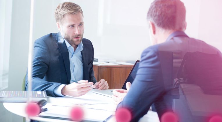 Two men in suits sit around a table and talk.