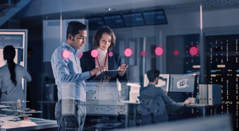 Several people can be seen in a futuristic office through a glass wall - two people in focus, looking at a tablet.