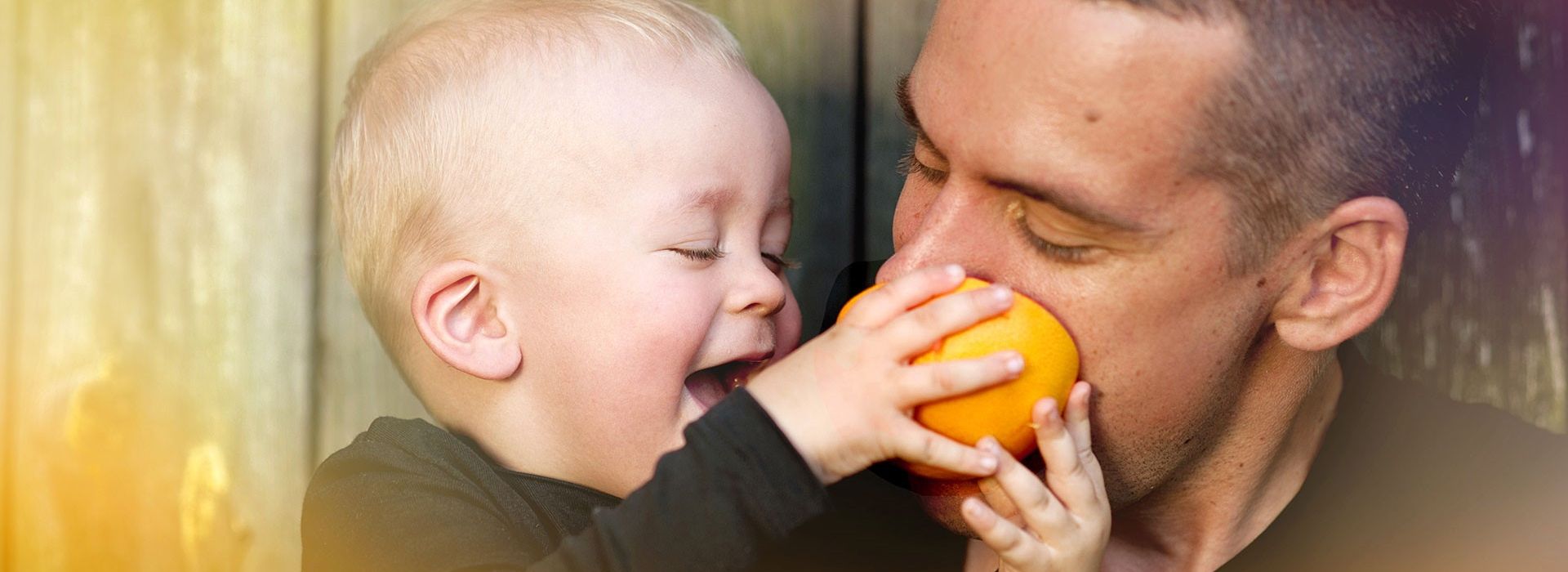A young man has a laughing child in his arms which makes him bite into an orange.
