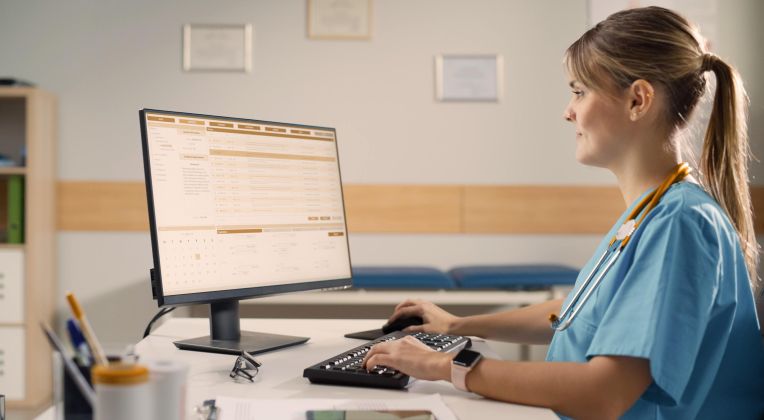 Junge Ärztin am Schreibtisch mit Stethoskop um den Hals arbeitet am Computer