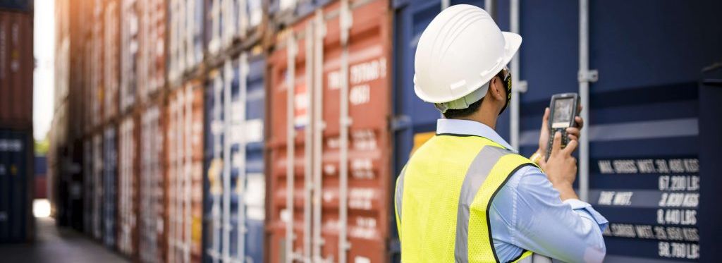 Ein Hafenarbeiter mit Warnweste und weißem Helm steht vor einer Containerwand und hält einen Scanner in der Hand.