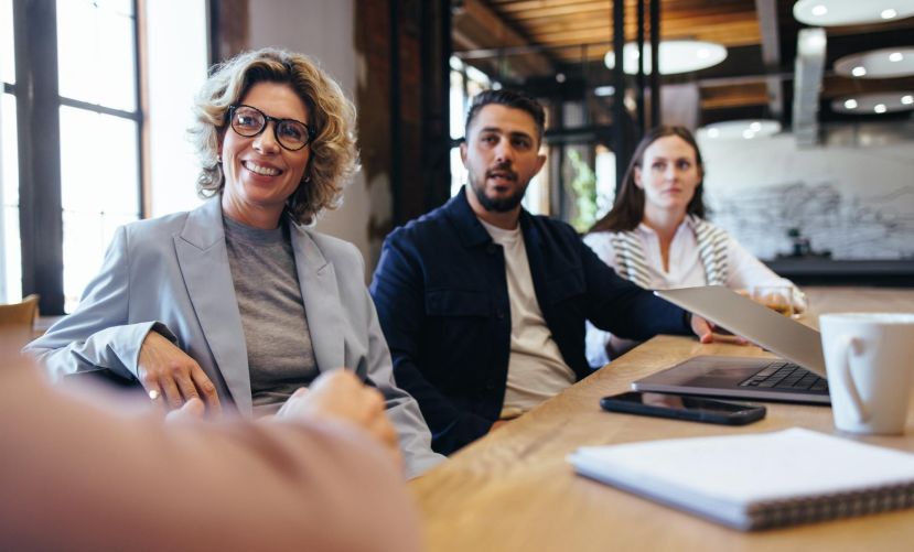 Junge Frau sitzt mit Kollegen am Tisch und schaut fröhlich über den Tisch.