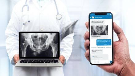 Ein Arzt hält ein Laptop in der Hand, auf dem ein Röntgenbild zu sehen ist. Auf einem Handy ist der LifeTime Messenger Chat zu sehen.
