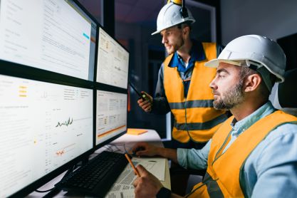 Two men in hard hats monitor machines on different monitors.