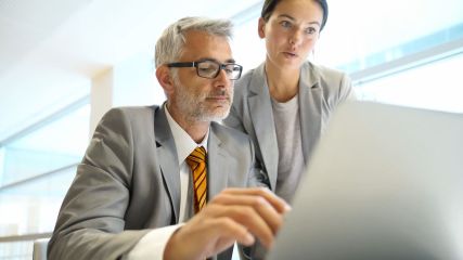 Ein Mann und eine Frau in Business Kleidung vor einem Laptop.