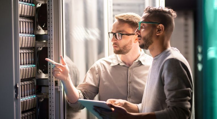 Two IT administrators stand in front of a rack and check data on a tablet.