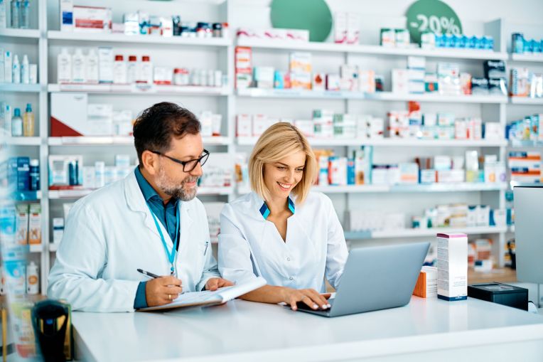 Zwei Apotheker: innen sitzen an einem Tisch in einer Apotheke und gucken auf einen Laptop