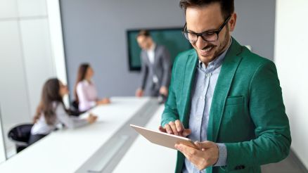 A man looks at data on a tablet in a conference room while a team of colleagues consults in the background.