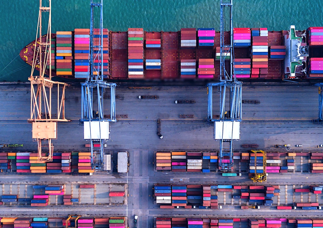 Bird's-eye view of the terminal. Four cranes including a ship with many containers. Trucks go back and forth.
