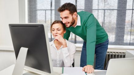 A woman sits in front of a PC and a man looks over her shoulder.