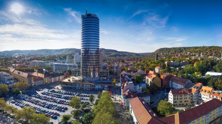 Skyline von der Stadt Jena, wo AKQUINET ebenfalls vertreten ist. Hochhaus, Häuser mit roten Dächern, Parkplatz, Bäume, hügelige Landschaft.
