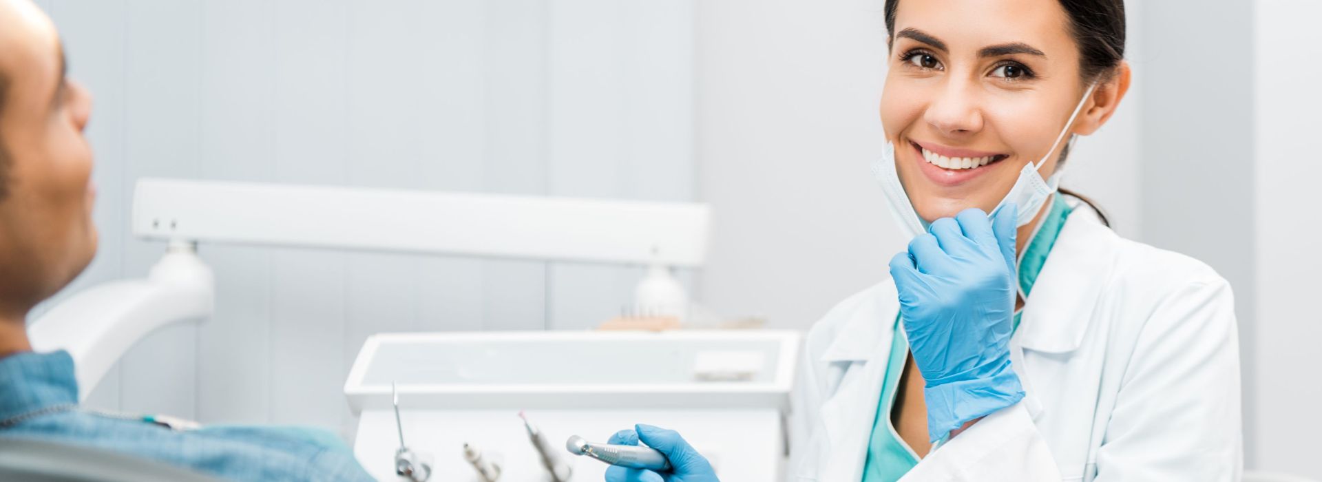 Eine Junge Zahnärztin lächelt in die Kamera und sitzt neben einem Patienten auf einen Behandlungsstuhl