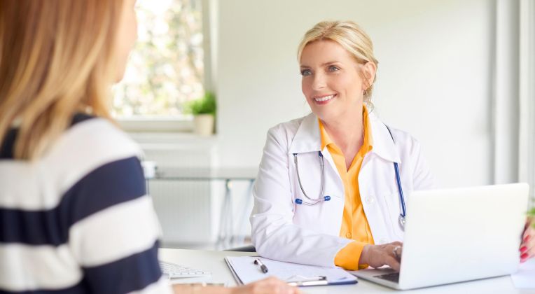 Ärztin sitzt am Schreibtisch vor einem Laptop und ist im Gespräch mit einer Patienten