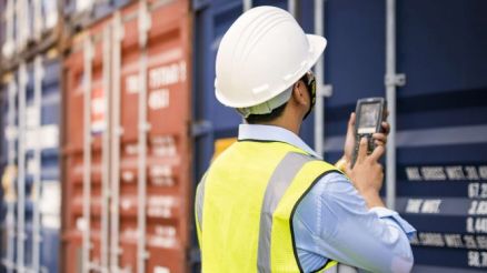 A port worker in a safety vest and white helmet stands in front of a container wall and holds a scanner in his hand.