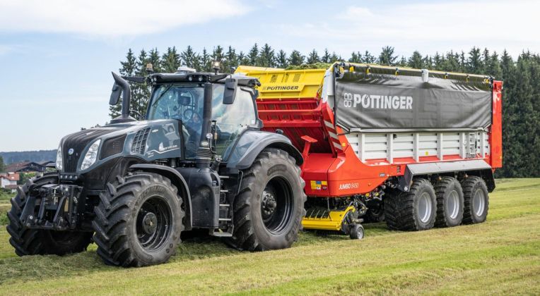 Modern black tractor with large PÖTTINGER JUMBO 8450 loader wagon on a freshly mown field with forest in the background.