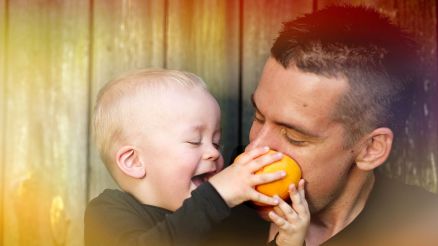 Ein junger Mann hat ein lachendes Kind auf dem Arm welches Ihn in eine Orange beissen lässt.