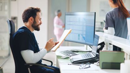 Ein junger Pfleger sitzt an einem Rechner in einem Krankenhaus und nutzt das Praxisverwaltungssystem LifeTime Cloud von AKQUINET.e