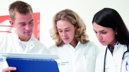 Krankenpflegerin lächelt mit verschränkten Armen in die Kamera. Hinter ihr, leicht verschwommen, diskutieren Arzt und weitere Helfer*innen im Gang eines Krankhauses.