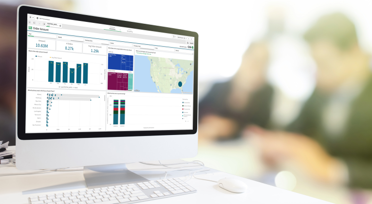 Während sich zwei AKQUINET Berater im Hintergrund unterhalten, steht ein Computerbildschirm mit Qlik Dashboard im Vordergrund. Es wird die Jahresansicht angezeigt mit verschiedenen Diagrammen von beispielsweise Geschäftsfeld und die Betragsentwicklung.