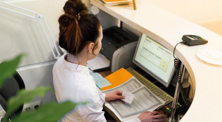 Eine Frau sitzt am Empfangstresen einer Arztpraxis und arbeitet am Computer