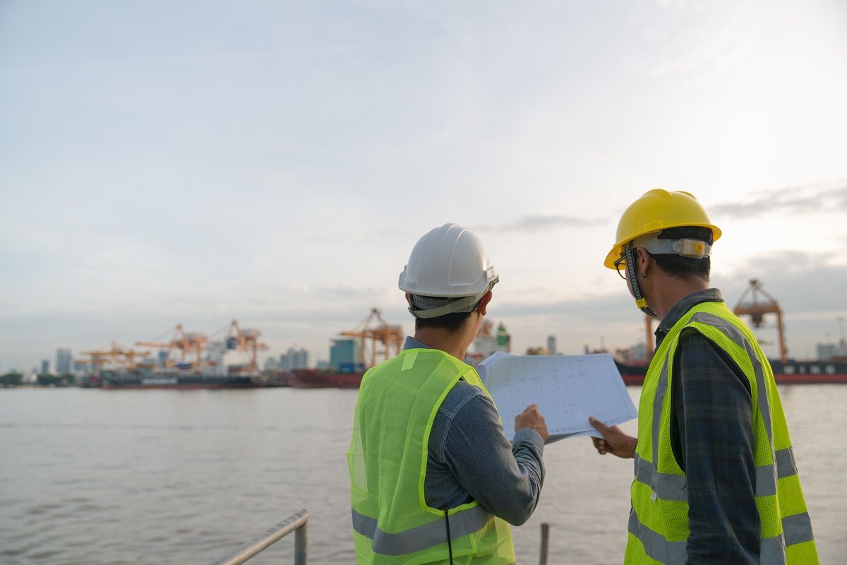Zwei Hafenspezialisten mit Warnwesten und Helmen, die ihre Planung in der Hand halten und über das Wasser zum Terminal schauen.