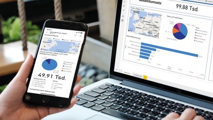 Eine Person sitzt vor einem Laptop, schaut sich ein Power BI Dashboard auf dem Monitor an und vergleicht es mit den Ergebnissen auf einem Handy.