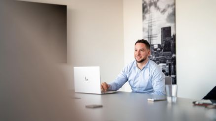 Ein Mann im blauen Hemd sitzt an einem Konferenztisch vor einem Laptop.