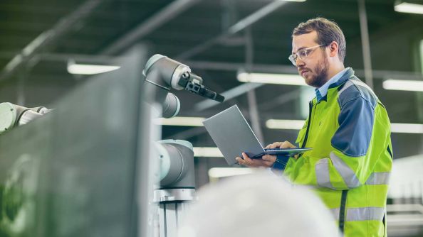 Ein Man mit gelb-grüner Sicherheitsjacke arbeitet mit einem Laptop in der Hand an einem Roboter