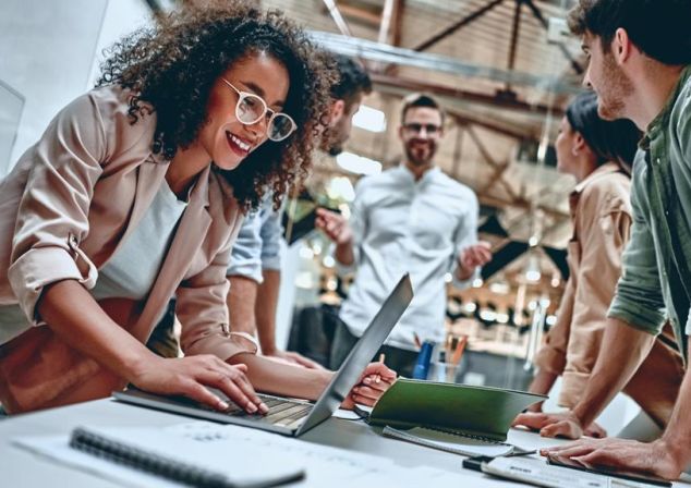 Eine Frau lächelt und beugt sich über ein Notebook, während sie mit vier Kolleg*innen an einem Projekt arbeitet. Mit Microsoft Fabric wird die teamübergreifende Zusammenarbeit durch den zentralen Zugriff auf alle benötigten Informationen gefördert.