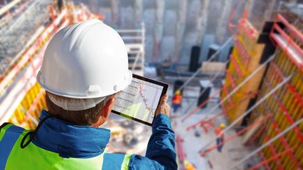 Ein Bauarbeiter schaut auf ein Tablet und steht dabei auf einer Baustelle.