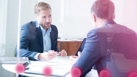 Zwei Männer im Anzug sitzen um einen Tisch und sprechen.