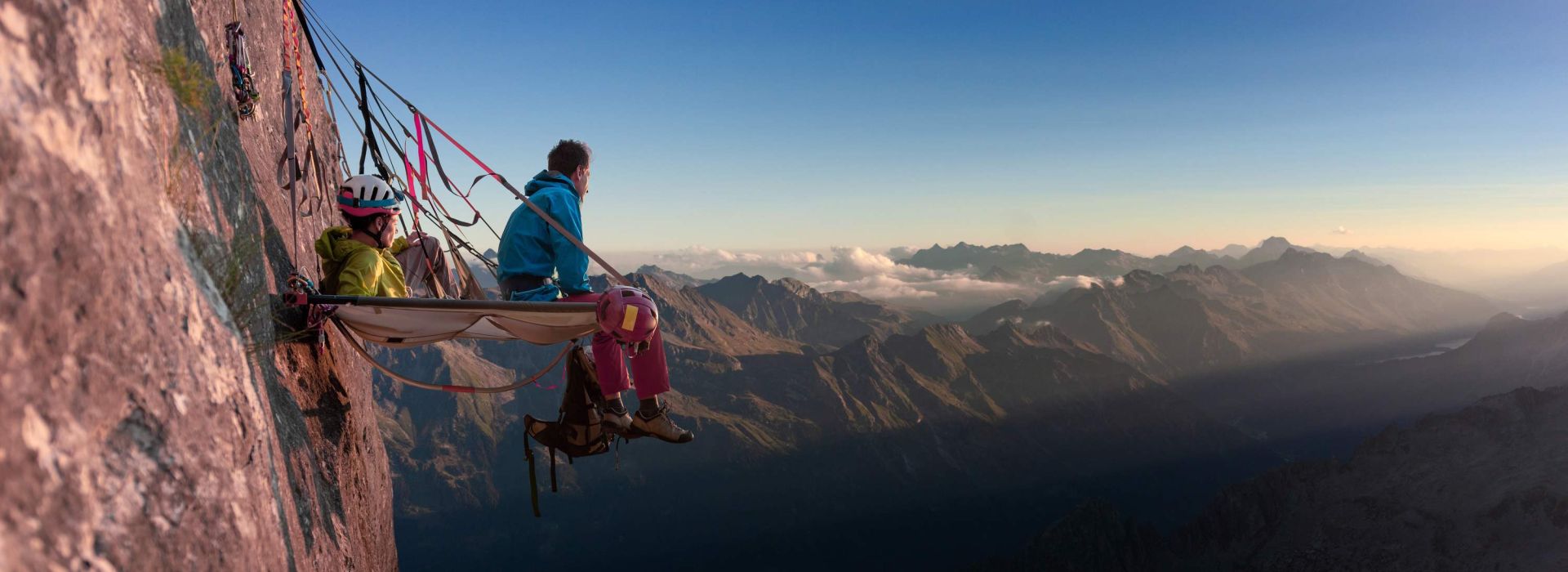 Zwei Personen sitzen auf einer Aussichtsplattform die an einer steilen Felswand befestigt ist und schauen auf eine Bergwelt.