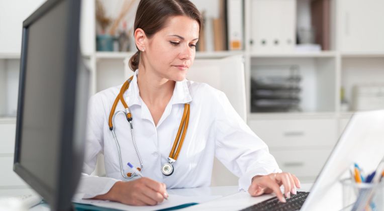 Ärztin mit weißem Kittel sitzt am Schreibtisch und tippt auf einem Laptop