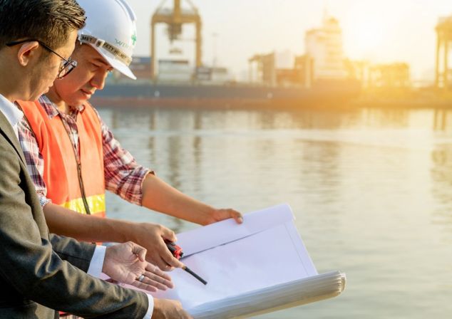 Dock worker and port consultant look at a folder. In the background, cranes of a port terminal.