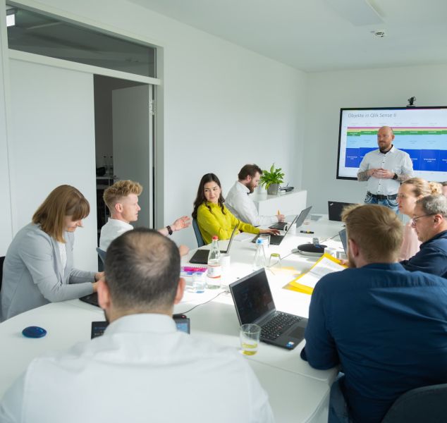 Schulungsteilnehmer mit Laptops unterhalten sich am Tisch, Trainer steht am Smartboard.