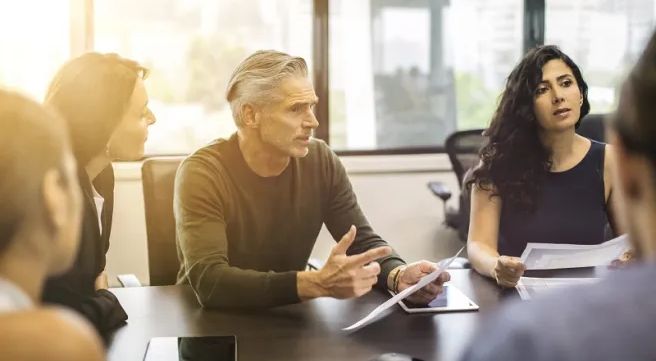 Several people sit at a table in a meeting and a man in the middle is talking.