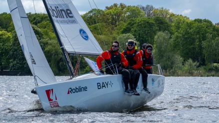 Weißes Segelboot mit vier Segler*innen mit schwarzen Hosen und roten Jacken auf einem See