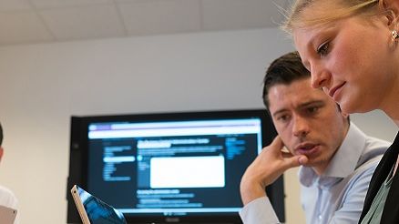 Zwei Kollegen besprechen etwas zu einem Microsoft Projekt und die Mitarbeiterin notiert sich etwas auf ihrem PC.