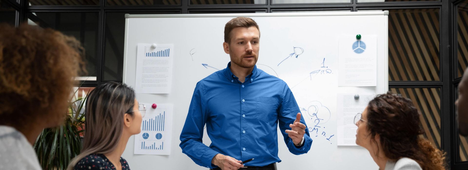 Ein Mann hält an einem Whiteboard eine Präsentation vor einem Team an Kolleg*innen.