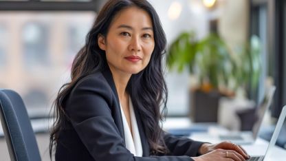 Eine junge Frau im Business Anzug sitzt am Computer und schaut in die Kamera