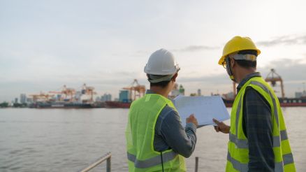 Two port consultants in high-visibility vests and helmets, holding their plan in their hands and looking across the water to the terminal.