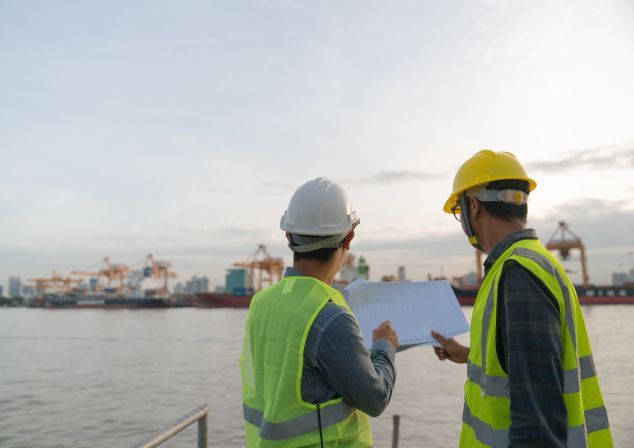Zwei Hafenspezialisten mit Warnwesten und Helmen, die ihre Planung in der Hand halten und über das Wasser zum Terminal schauen.