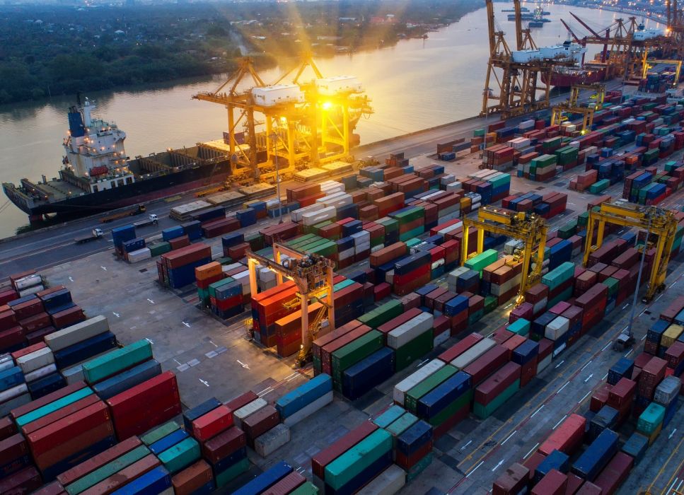 Hafenterminal bei Sonnenuntergang mit riesigem Containerschiff, Kränen und vielen verschiedenen Containern.