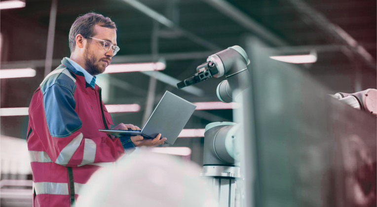 Ein Ingenieur in einer Industriehalle arbeitet mit einem Laptop an einer Robotik-Anlage.