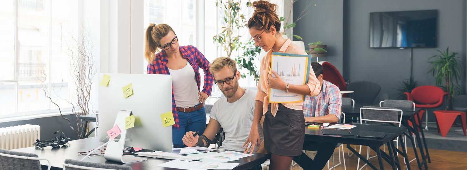 Drei Teammitglieder beraten sich zur Datenintegration an einem Schreibtisch mit großem PC-Bildschirm und schauen sich Unterlagen dazu an.