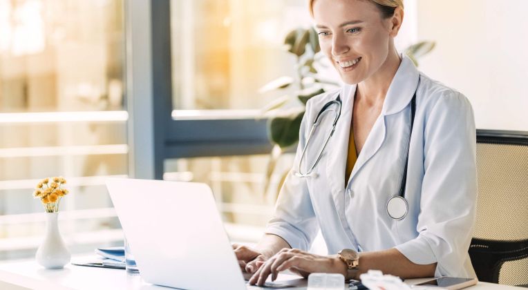 Eine Ärztin mit Stethoskop um den Hals sitzt am Schreibtisch und arbeitet am Laptop